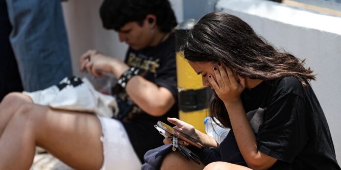 Two young people sit outdoors, one looking at a phone with hands on cheeks, the other blurred in background.