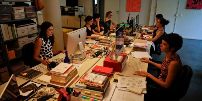 A row of office workers seated at a long desk with laptops and monitors, surrounded by books and papers on a cluttered workspace.