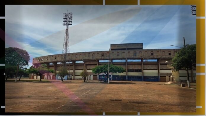 Front view of a large concrete stadium with a tall floodlight tower and empty plaza under a blue sky, trees lining the foreground.