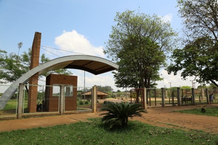 Entrance to a park with a curved white arch over a gate, brick pillars, and a chain-link fence nearby.