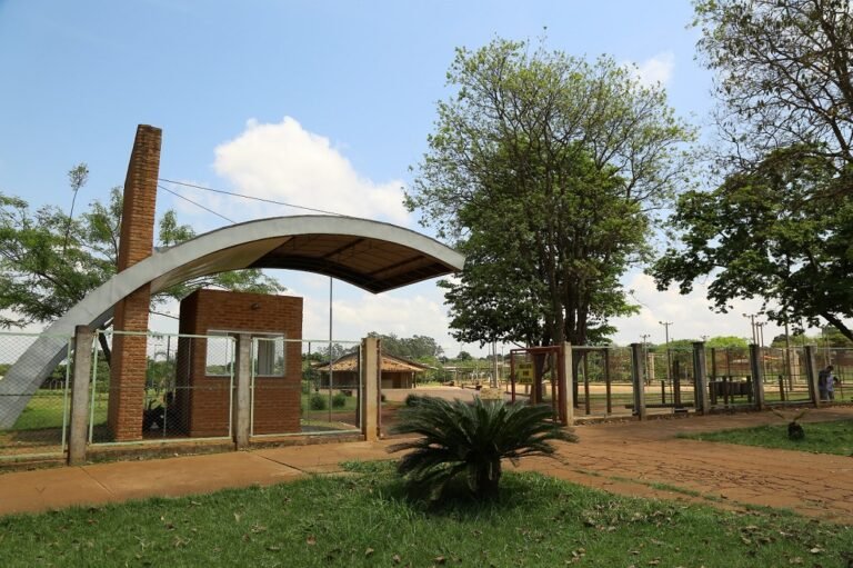 Entrance to a park with a curved white arch over a gate, brick pillars, and a chain-link fence nearby.