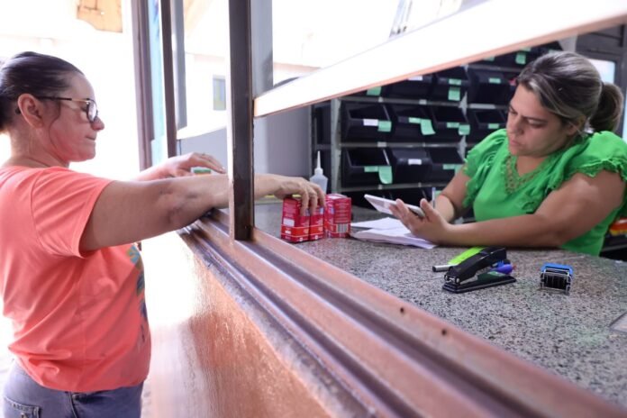 Customer in a pink shirt hands items to a clerk at a window service counter; countertop with small red boxes nearby.