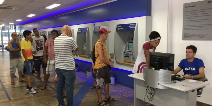 Queue of diverse customers waiting at ATMs in a bank lobby with a teller at a desk nearby.