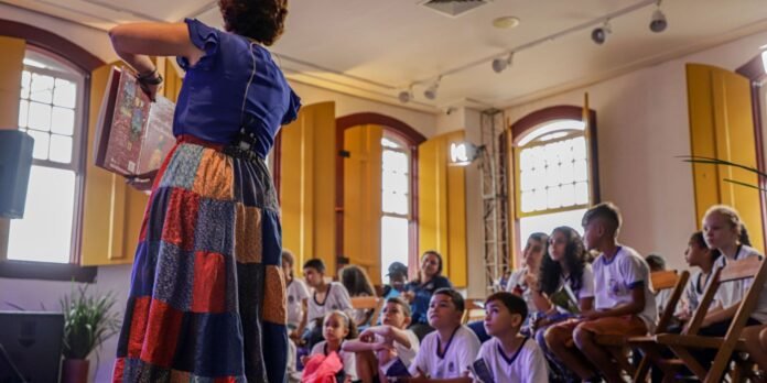 Storyteller in a blue top and patchwork skirt reads from a book to children sitting in a bright classroom.