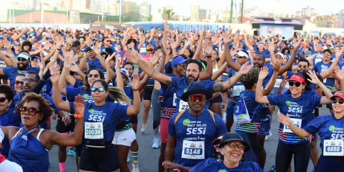 Large crowd of runners in blue SESI shirts raise arms at a city race start, smiling and energized.