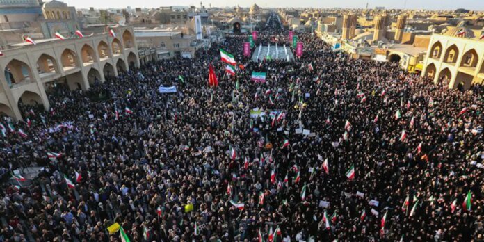Overhead view of a dense crowd in a square lined with arches, many people waving green, white, and red Iranian flags and banners during a protest.