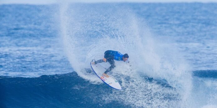 Male surfer in a blue shirt rides a breaking wave, spray shooting up as he carves with a white board.