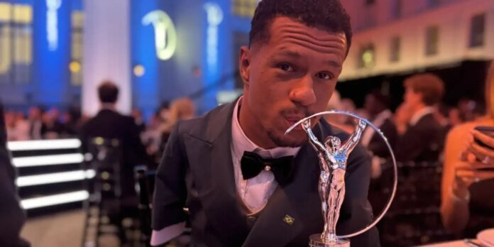 Man in a black tuxedo and bow tie kisses a silver award statue with a circular ring at a formal event.