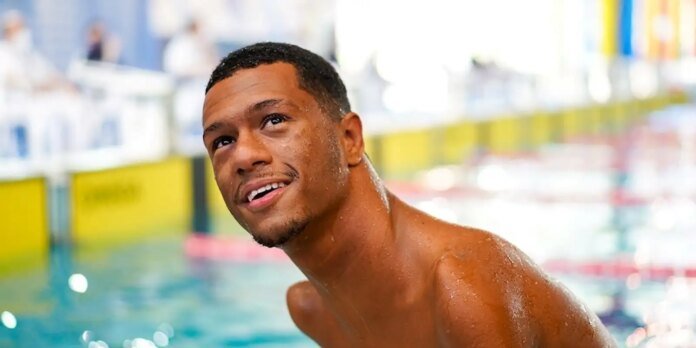 Smiling young man with wet skin at an indoor swimming pool, leaning slightly toward the camera.