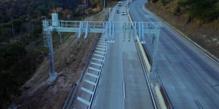 Aerial view of a highway with a large overhead gantry and road markings, showing a construction zone on the left shoulder and a few cars in the distance.