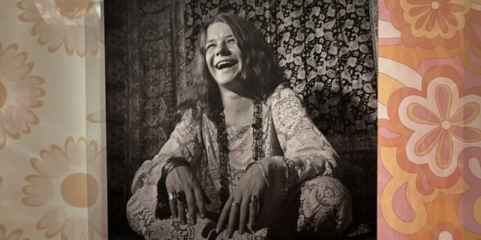 Laughing woman in a lace bohemian outfit sits against a patterned tapestry, flanked by vintage floral curtains (black-and-white photo).