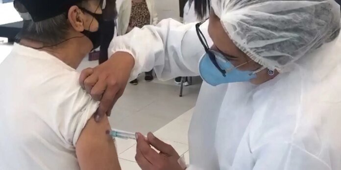 Healthcare worker in PPE giving a vaccine to a person’s upper arm while both wear masks; injection in progress in a clinical setting.