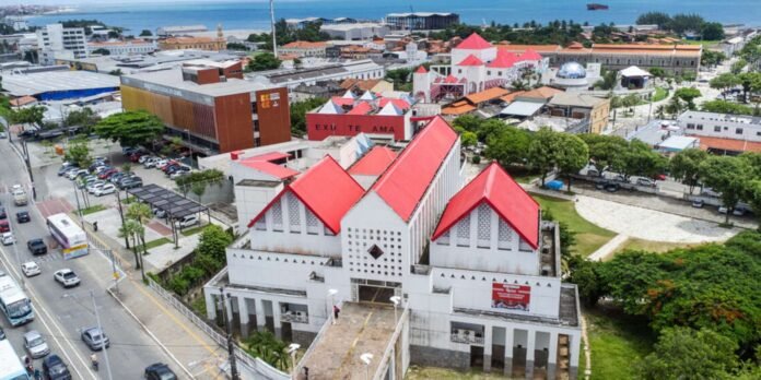 Aerial view of a coastal town showing a white building with three bright red pitched roofs among red-tiled rooftops and trees by the street.