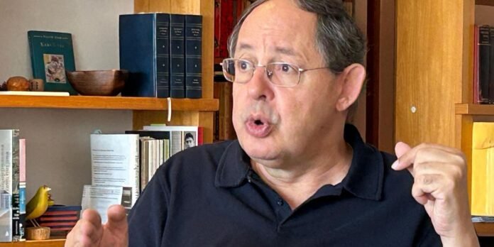 Man wearing glasses and a dark polo speaks, gesturing with his left hand in a library or study setting.