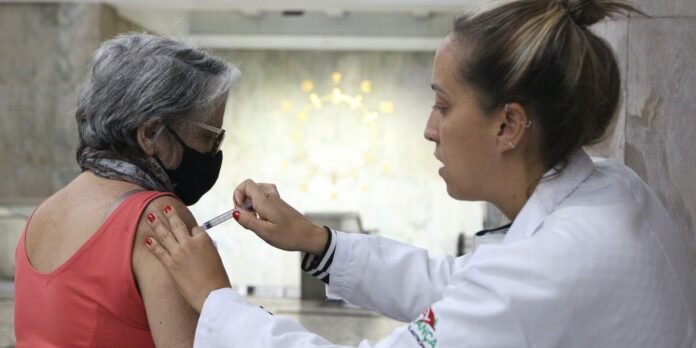 Nurse in a white coat administers a vaccination to an older woman wearing a pink top and black mask in a clinical setting.