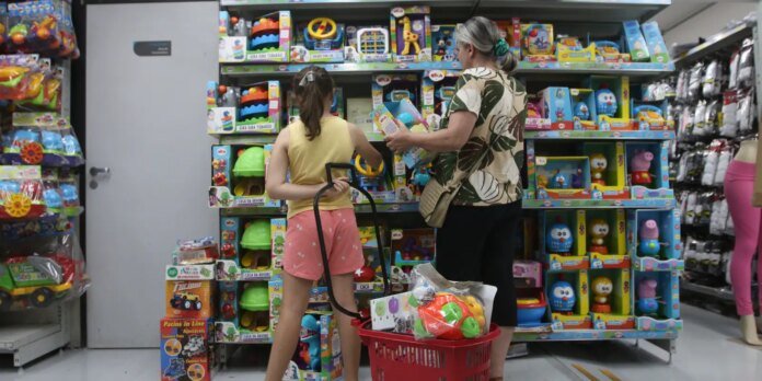 Mother and daughter browse toy shelves in a colorful store aisle filled with children's toys and boxes.