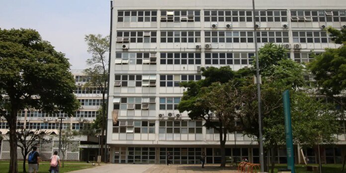 Multi-story gray office building with many windows and wall-mounted AC units, with green trees and pedestrians in front.
