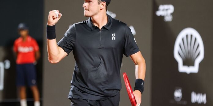 Male tennis player in a black polo celebrates with a clenched fist, holding a red racket on the court.