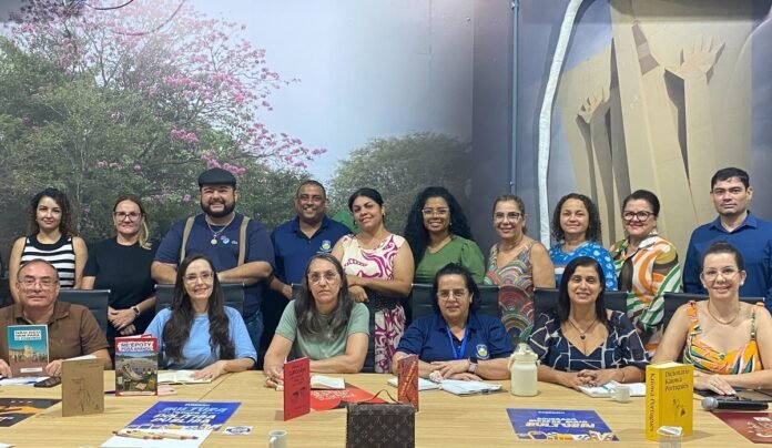 Group of diverse adults posing around a conference table with books and notebooks, in a classroom or meeting room setting.