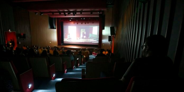 Dimly lit theater with red stage curtains and an audience watching a presentation on a bright, projected screen at the front.
