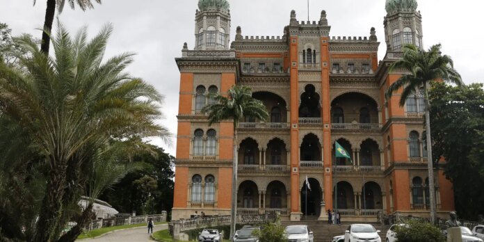Ornate colonial-style orange and beige building with arched balconies and two towers, palm trees in front, Brazil flag visible.