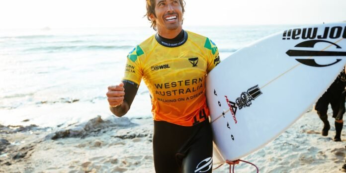 Surfer in a bright yellow Western Australia jersey walks along a sunlit beach, carrying a white surfboard under his arm.