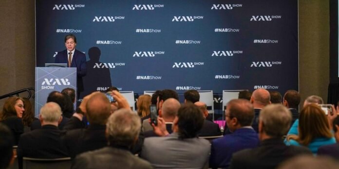 Male speaker in a navy suit at a podium presenting at the NAB Show, blue backdrop covered with NAB Show logos and an attentive audience in front.