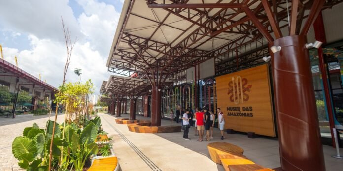 Group of people standing under a covered walkway at Museu Das Amazonas, with wooden sign and modern columns nearby.
