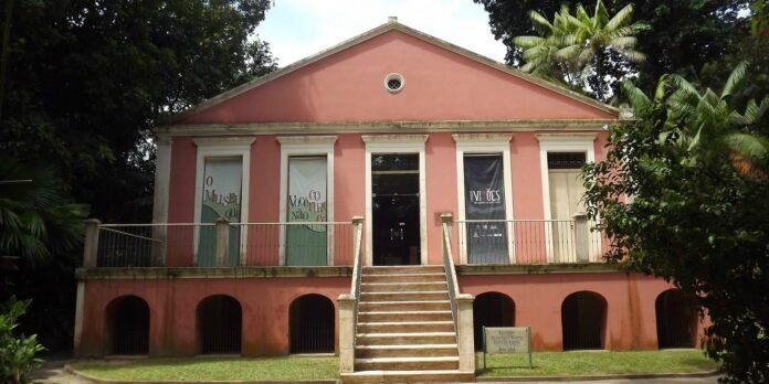 Pink colonial-style museum building with a central staircase, white pillars, and banners in tall ground-floor windows set among tropical trees.