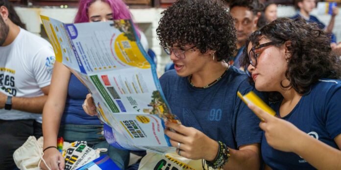 Two young people closely inspect a large, colorful brochure in a crowded setting.