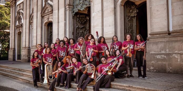A large group of young women in maroon shirts posing with brass and string instruments on church steps outside a stone building.