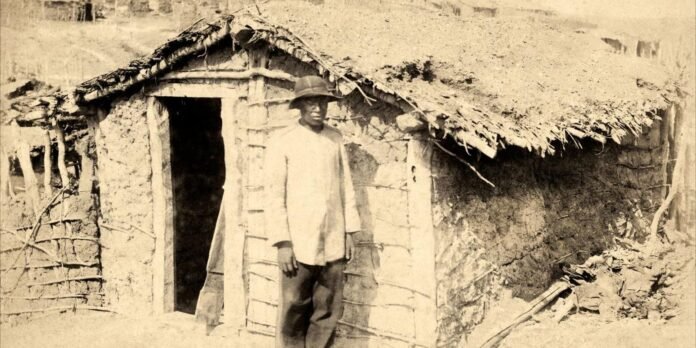 A man in a light shirt and hat stands outside a small mud-brick hut with a thatched roof in a rural setting.