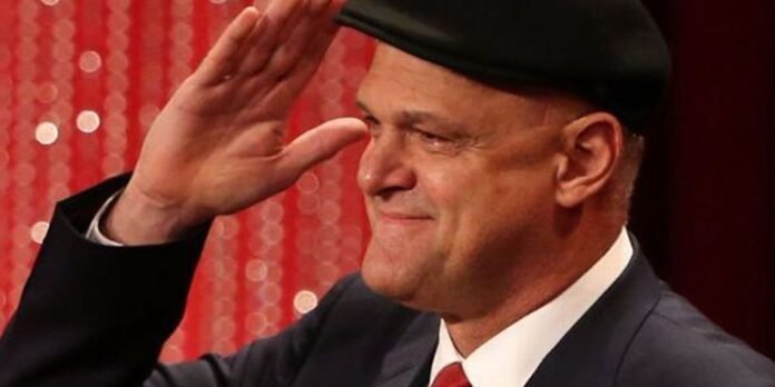 Man in a dark suit with a red tie tipping a black fedora on a red sequin backdrop.