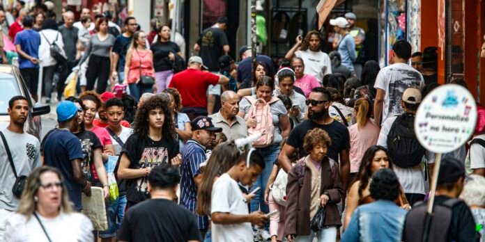 Crowded city sidewalk with diverse pedestrians walking and talking, storefronts visible in the background.