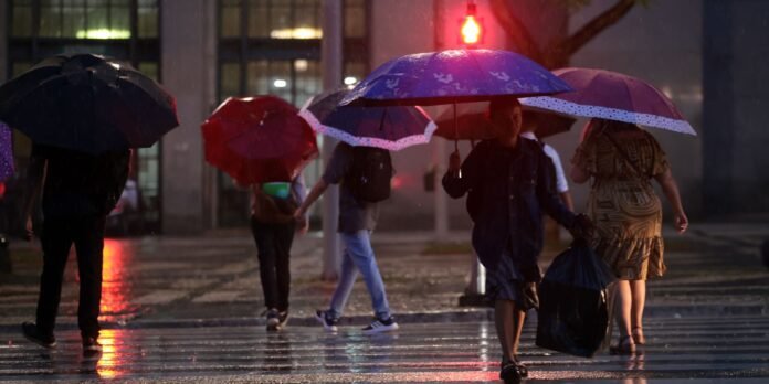 People walking in the rain with colorful umbrellas on a wet city street at night, reflections on the pavement visible behind them.