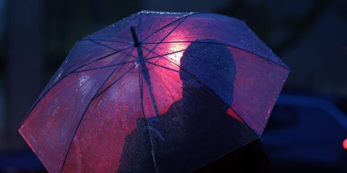 Person under a red umbrella in the rain at night, lit by blue neon light with raindrops on the canopy.