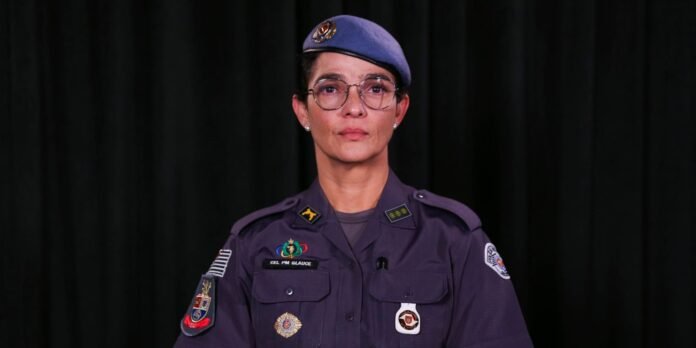 Police officer in a purple uniform and beret, standing against a dark curtain, with badges on the chest and sleeve.