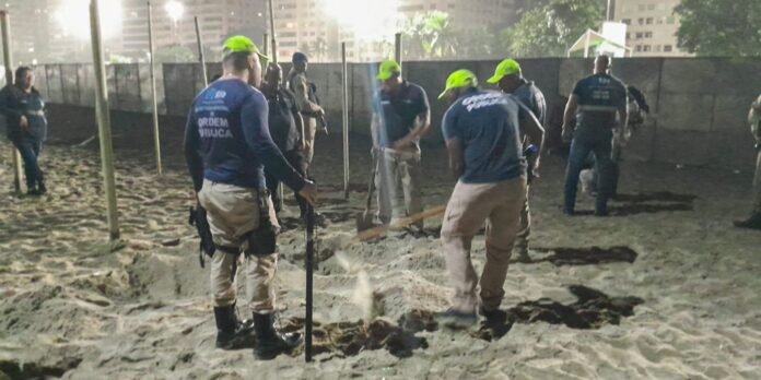 Group of uniformed workers wearing blue shirts and neon green helmets standing around a shallow trench on a sandy lot at night, with one person holding a shovel.