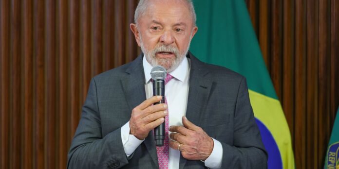Older man in a gray suit speaks into a handheld microphone at a formal event, with a Brazilian flag in the background.