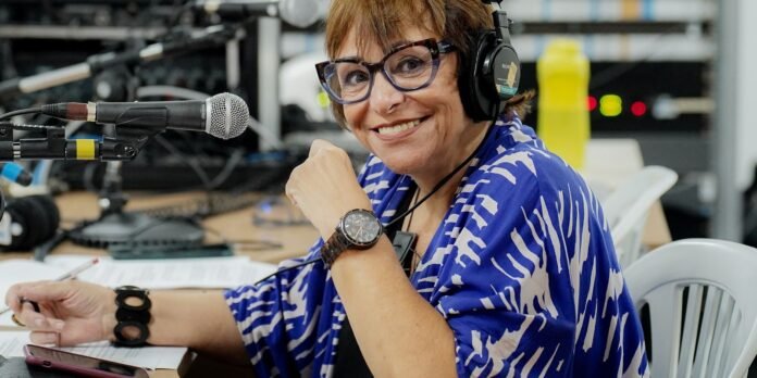 Smiling woman wearing headphones and a blue patterned blouse speaks into a microphone in a recording studio with papers and equipment nearby