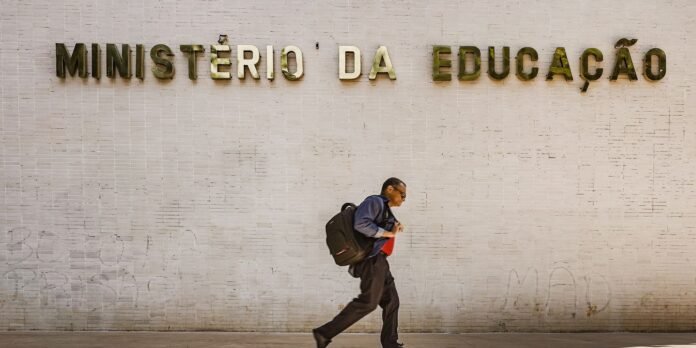 Man with a large black backpack walks briskly past a pale brick wall with the sign 'MINISTÉRIO DA EDUCAÇÃO' in Portuguese.