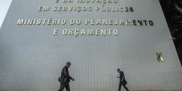 Two men walk in front of a large tiled wall with a gold-lettered sign reading 'MINISTÉRIO DO PLANEJAMENTO E ORÇAMENTO'.