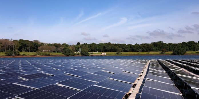Extensive solar panel arrays on floating platforms across a calm body of water, with a tree-lined shore and blue sky behind.
