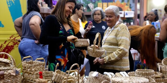 Smiling woman in a floral dress hands a small woven basket to an elderly vendor at a busy market stall full of baskets.