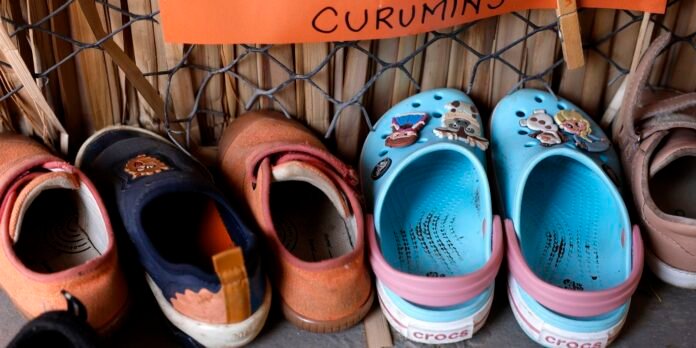 Row of children's shoes on a shelf, including orange sneakers, a dark blue shoe, tan sandals, and blue Crocs decorated with cartoon pins.