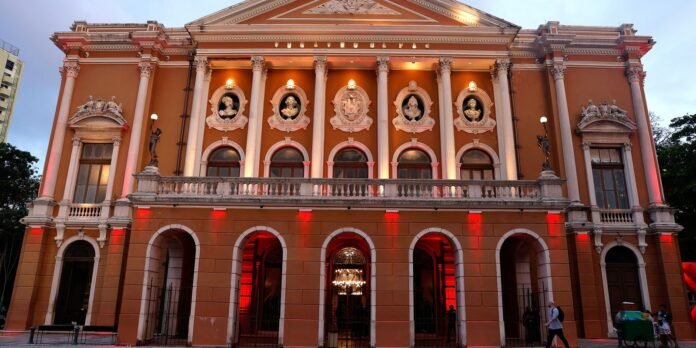 Ornate orange neoclassical building with white columns and arched entrances; statues along the balcony balustrade and a chandelier visible through the central arch.