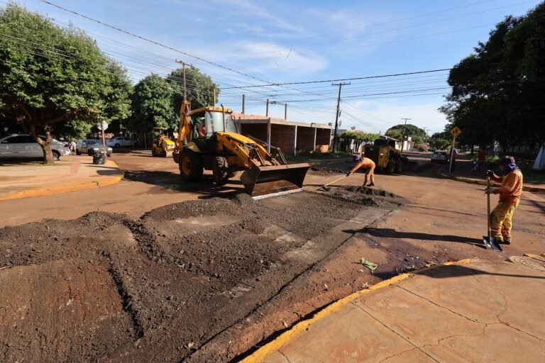 Construction crew repairs a street with a yellow backhoe and workers spreading asphalt, dirt piled in front. (informative)