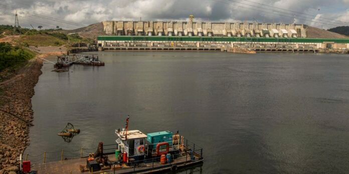 Industrial dam with multiple spillways across the top, green maintenance building at waterline, and a calm reservoir in the foreground; a work barge floats near a rocky shoreline on the left.