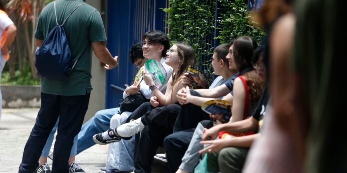 Group of young people sitting on a curb, listening to a man with a blue drawstring backpack standing and talking to them at an outdoor event.