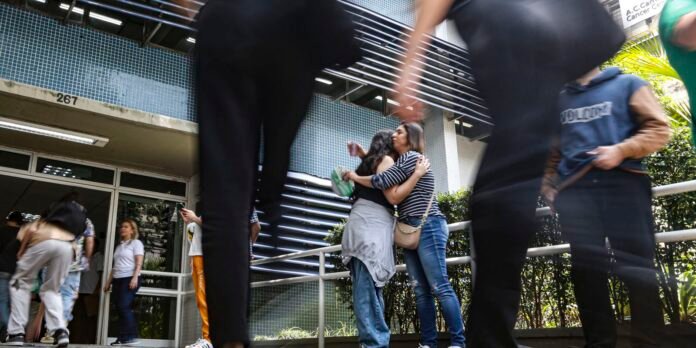 Two women share a hug near a railing outside a blue-tiled building entrance while others walk by. Building number 267 is visible above the doorway.